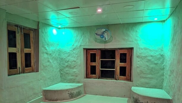Una habitación con techo iluminado en verde, un ventilador de techo y dos ventanas con marcos de madera.
