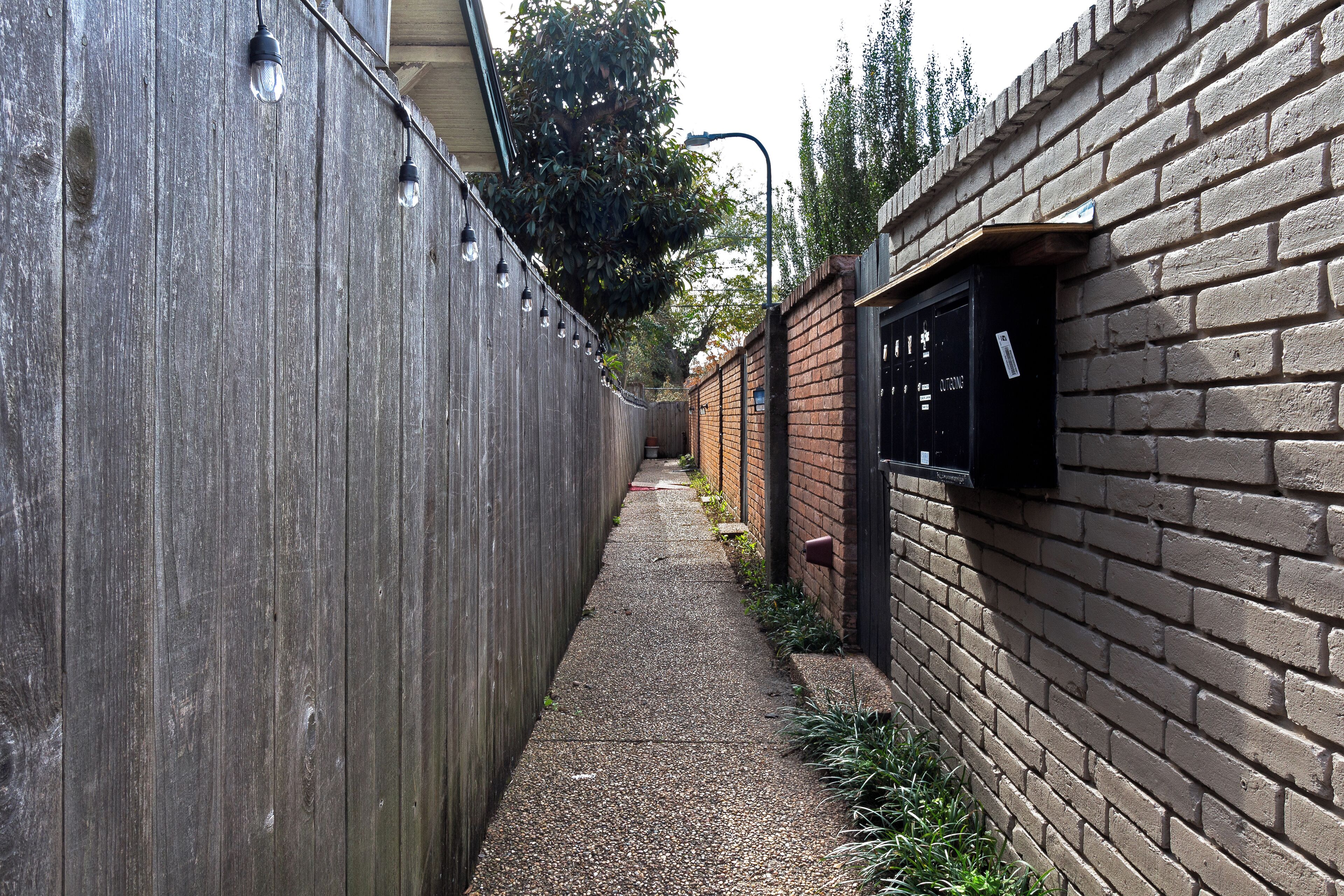 A narrow alleyway with a wooden fence on one side and a brick wall on the other, leading to a building entrance.