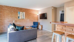 Un salon avec un mur en briques, un canapé gris, un fauteuil bleu et un comptoir de bar en bois avec des tabourets.