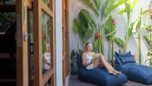 Eine Person entspannt auf einer blauen Außensofa am Pool, im Hintergrund üppige tropische Pflanzen und eine Holzterrasse.