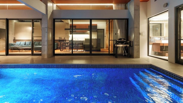 Una piscina interior moderna con paredes de vidrio transparente, techo de madera y un área de comedor visible a través del vidrio.