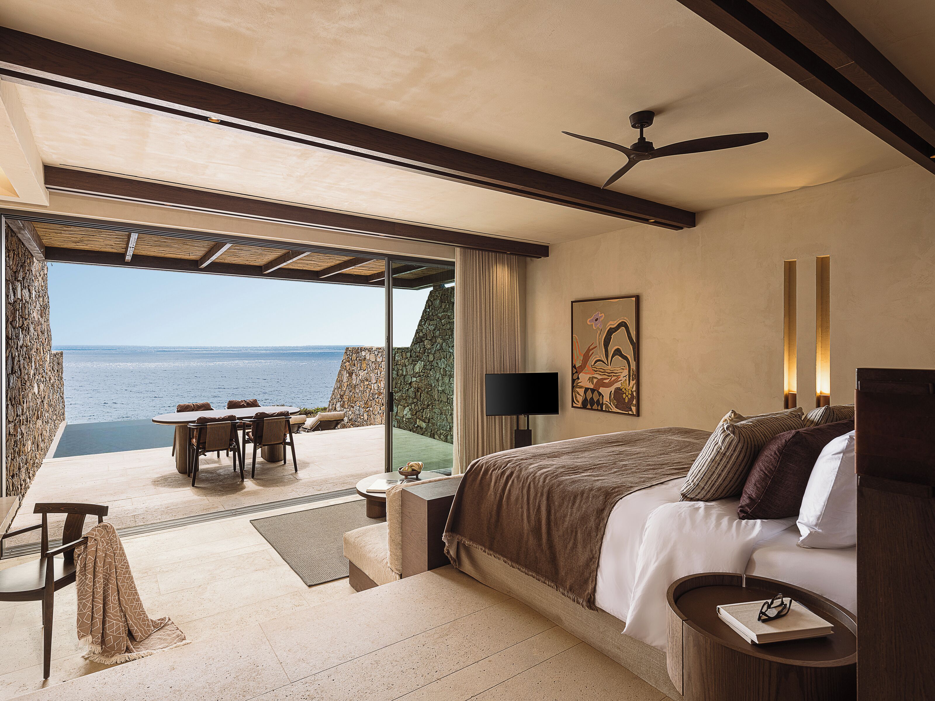 A bedroom with a bed, a desk, and a chair, a large window with a view of the sea, and a ceiling fan.
