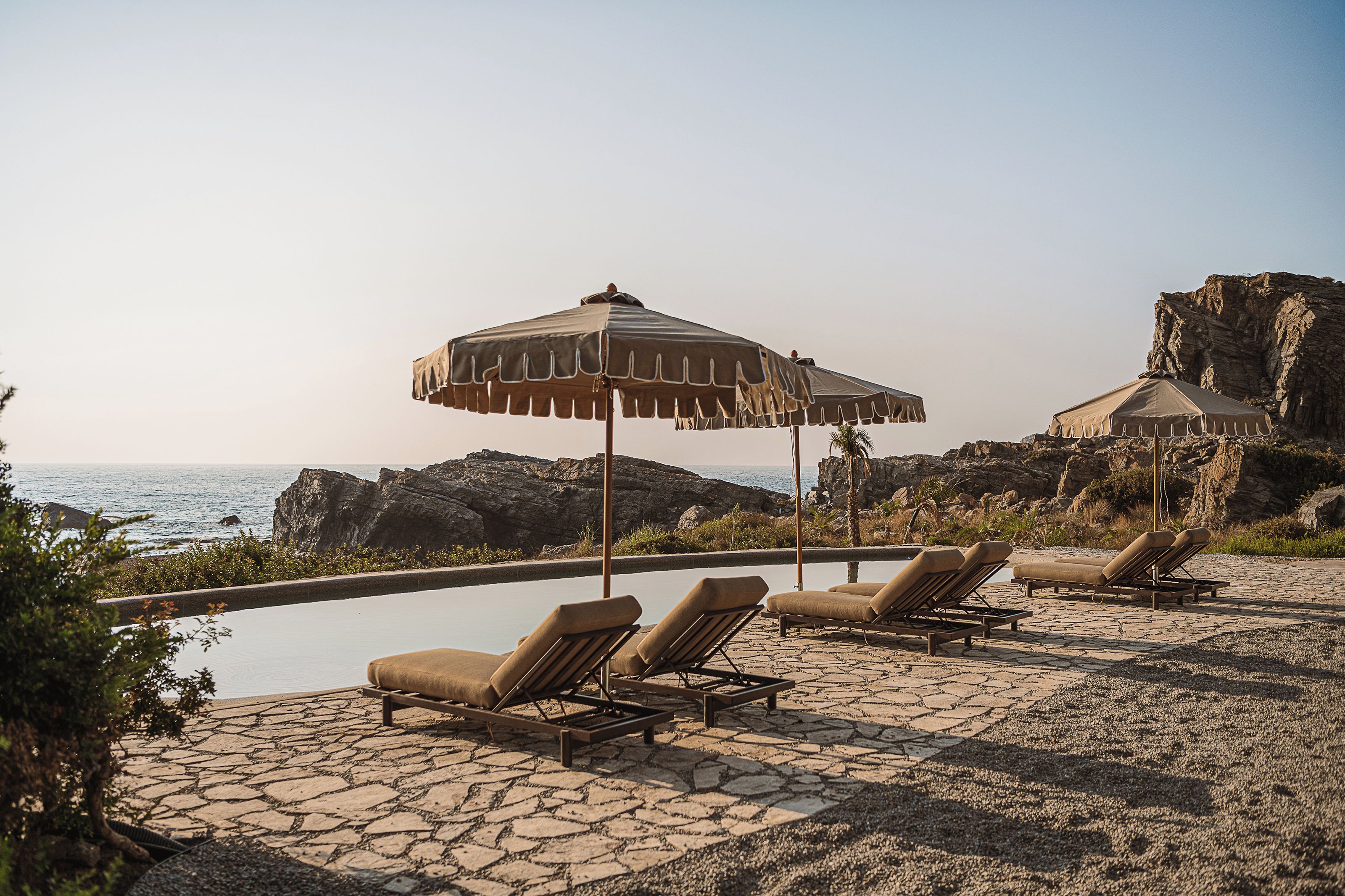 A poolside area with lounge chairs and umbrellas, overlooking a rocky coastline and the sea.