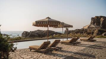 A poolside area with lounge chairs and umbrellas, overlooking a rocky coastline and the sea.