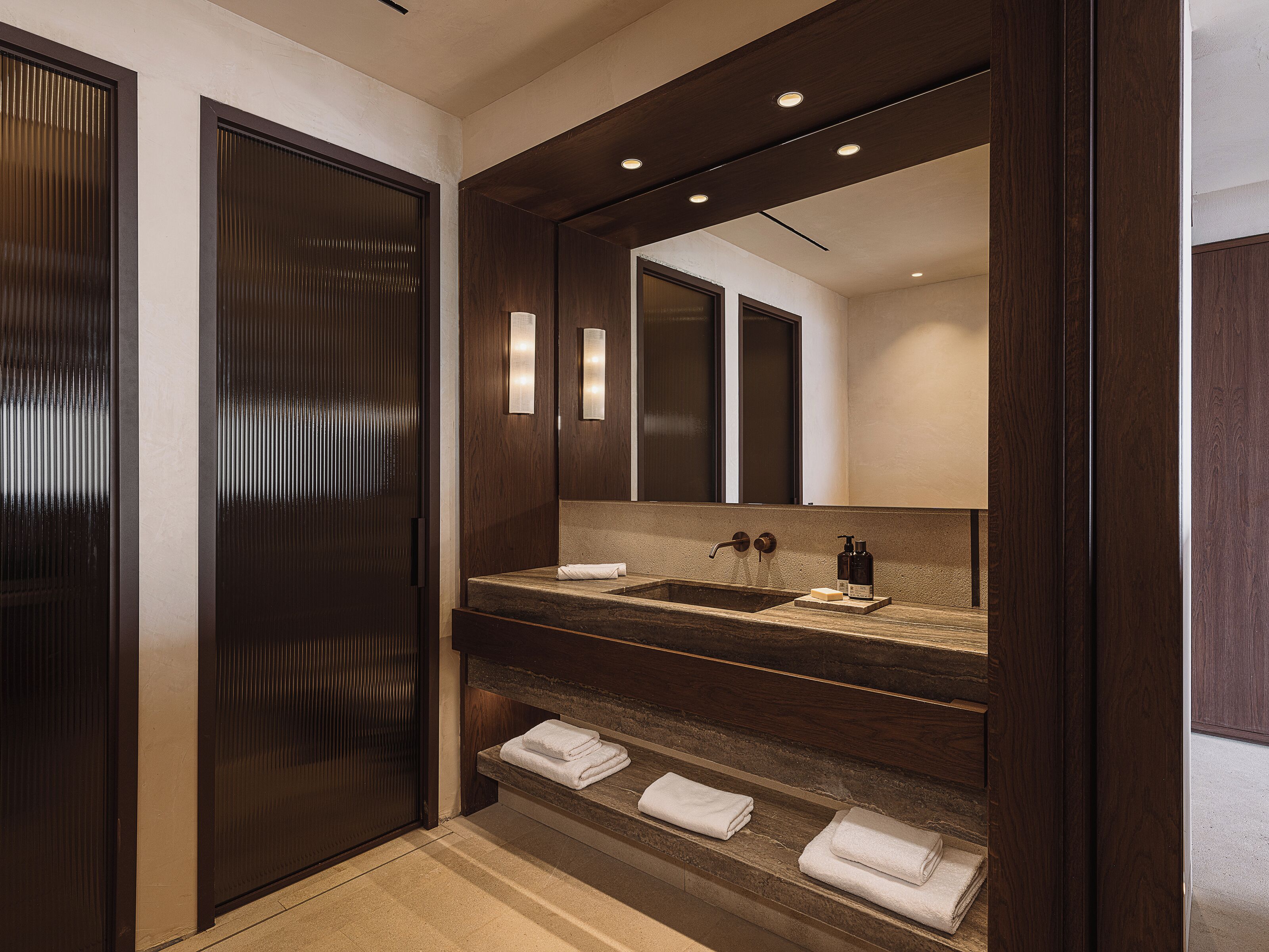 Une salle de bain moderne avec un meuble-lavabo en bois foncé, un grand miroir et une étagère intégrée où sont rangées les serviettes.