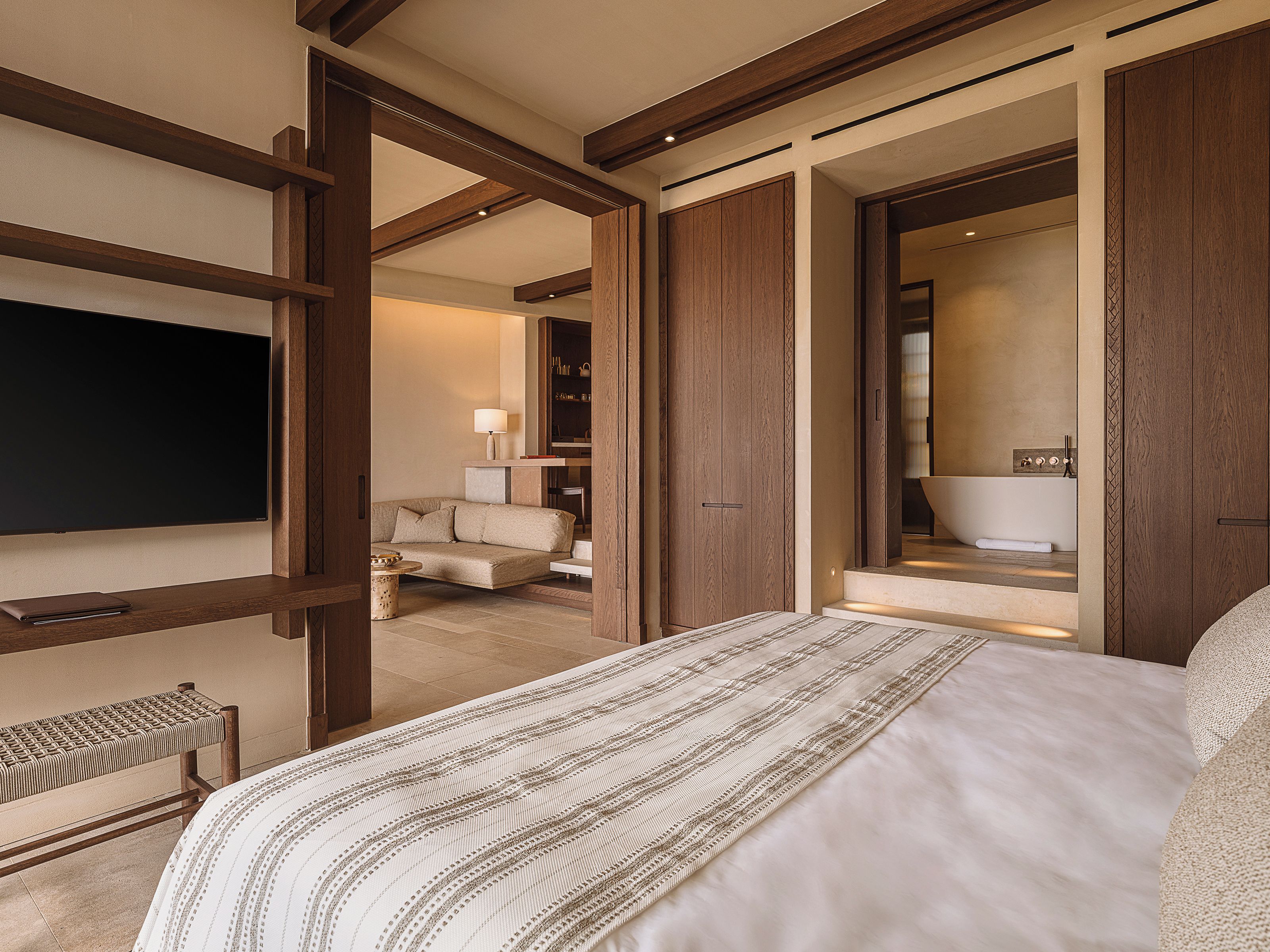 A hotel room with a large bed, a seating area with a sofa and armchairs, a flat-screen TV, and a bathroom with a bathtub and mirror.