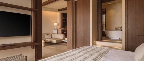 A hotel room with a large bed, a seating area with a sofa and armchairs, a flat-screen TV, and a bathroom with a bathtub and mirror.