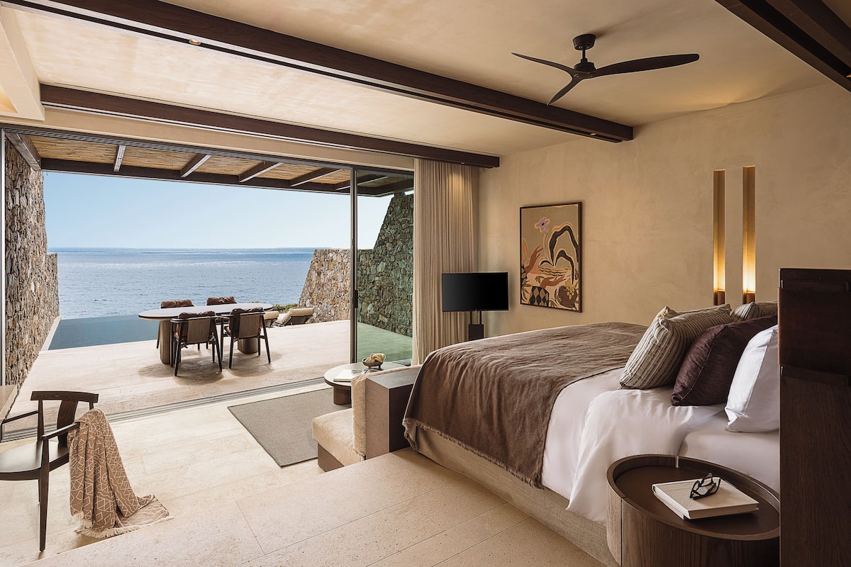 A bedroom with a bed, a desk, and a chair, a large window with a view of the sea, and a ceiling fan.