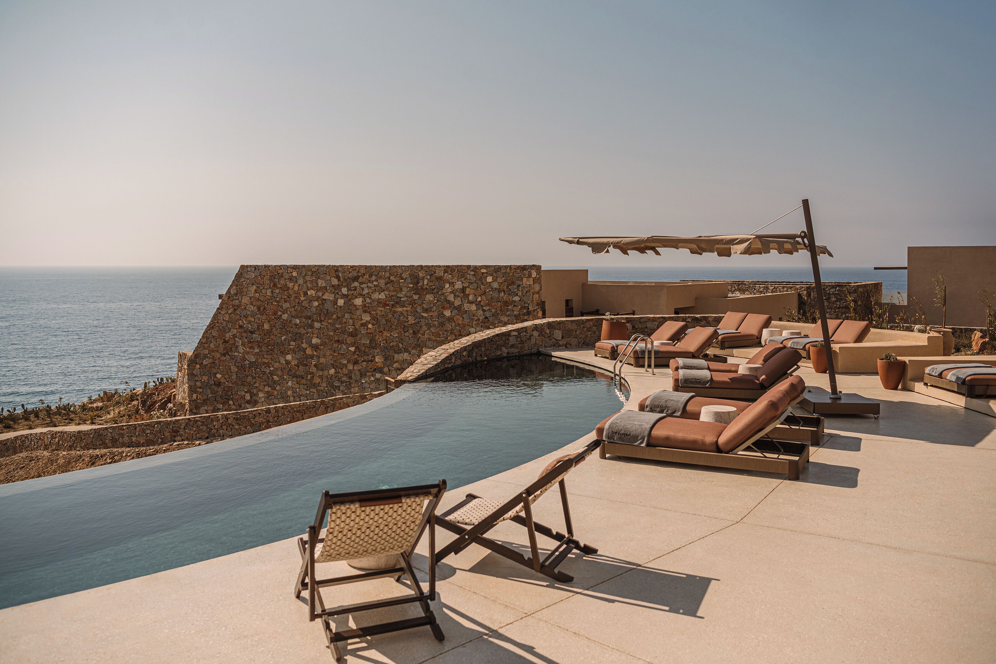 A rooftop infinity pool with lounge chairs and a stone wall overlooking the sea.