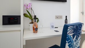 Een moderne hotelkamer met een wit bureau, een flatscreen televisie aan de muur, een potorkidee, een wijnglas, een fles wijn en een stoel met een kussen met tropisch dessin.