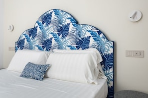 Un lit avec du linge de lit blanc et un coussin à motifs bleus, une tête de lit ornée de motifs de feuilles tropicales et un petit pouf rond.