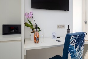 Une chambre d’hôtel moderne avec un bureau blanc, un téléviseur à écran plat fixé au mur, une orchidée en pot, un verre à vin, une bouteille de vin et une chaise avec un coussin à motifs tropicaux.