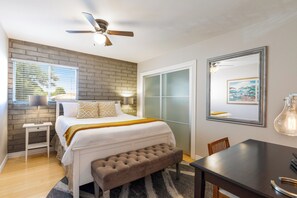 Un dormitorio con cama, ventilador de techo, ventana con persianas, mesita de noche, banco, mesa de comedor y un cuadro en la pared.