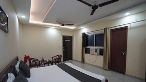 A hotel room with a bed, a ceiling fan, a window with curtains, and a small table with chairs. - NOVA RESORTS (Raebareli)