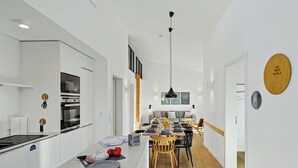 Eine moderne Küche mit einer zentralen Kochinsel, ein Essbereich mit einem Holztisch und Stühlen sowie ein Gang zu einem weiteren Raum.