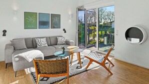 Ein modernes Wohnzimmer mit einer Couch, zwei Sesseln, einem Couchtisch und einem Teppich. An der Wand hängen drei gerahmte Bilder und ein Schiebetürenfenster führt in einen Außenbereich.