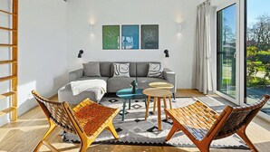 Ein modernes Wohnzimmer mit einem grauen Sofa, zwei Holzstühlen, einem kleinen runden Tisch und einem großen Fenster mit Blick auf Grünflächen.