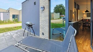 Een modern terras buiten met een barbecue, een glazen deur naar een eethoek en uitzicht op de omgeving.