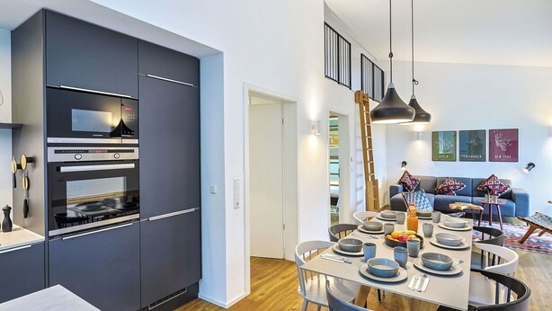 Een moderne keuken met inbouwapparatuur, een eethoek met een ronde tafel en stoelen, en een slaapverdieping bereikbaar via een ladder.