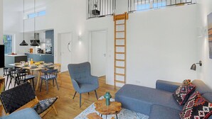 Un vaste espace de vie comprenant un coin repas, un canapé gris, une échelle en bois et une vue sur la cuisine.