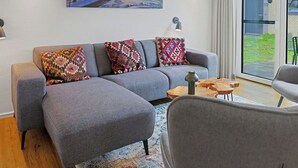 Un salon moderne avec un canapé gris, des coussins à motifs, une table basse en bois et une grande fenêtre donnant sur l’extérieur.
