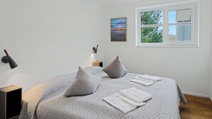 Une chambre à coucher avec un lit, des oreillers, une table de chevet, une lampe, une fenêtre et un tableau.