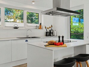 Une cuisine moderne dotée d’un îlot central, d’un évier et d’une cuisinière. De grandes fenêtres offrent une vue sur la verdure environnante.