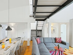 Ein modernes Interieur mit Essbereich, einem grauen Sofa mit gemusterten Kissen und einer Treppe, die in eine obere Ebene führt.