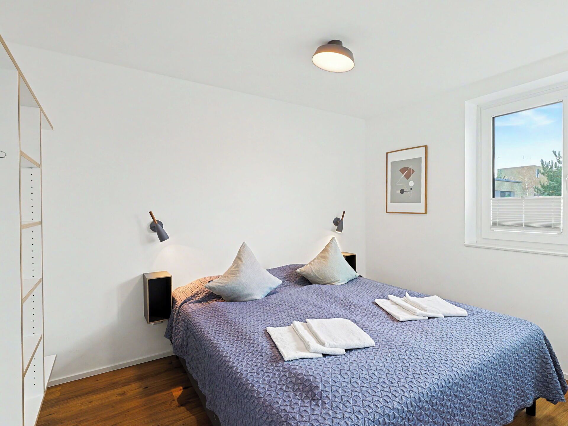 A bedroom with a bed, pillows, a wall-mounted light, a window, and a framed picture.