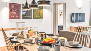 Ein moderner Essbereich mit einem Holztisch, gedeckt für vier Personen, einschließlich Schüsseln, Gläsern und einer Obstschale. Drei Pendelleuchten hängen darüber, und an der Wand ist Kunstwerk mit Stadtmotiven zu sehen.