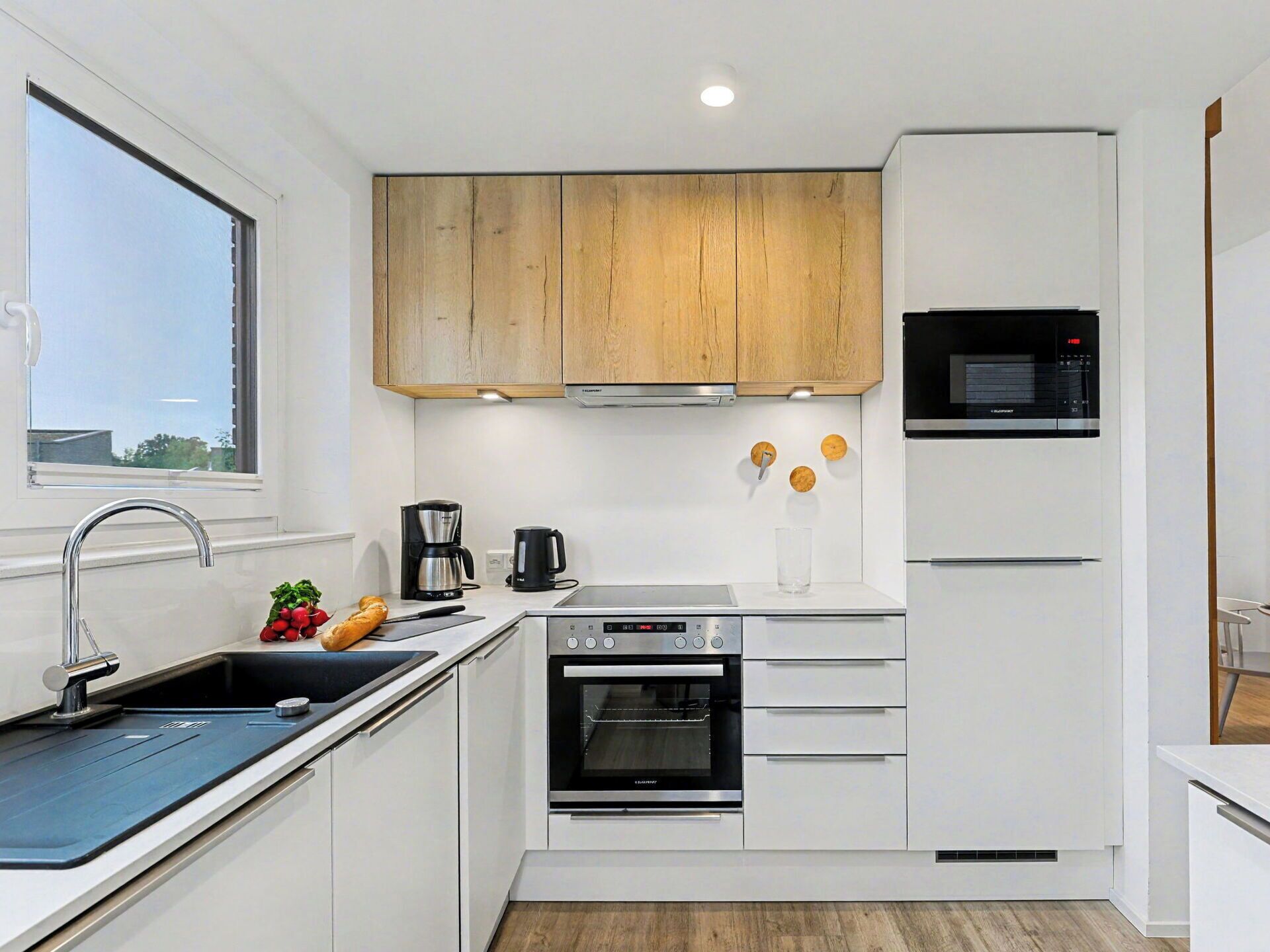 A modern kitchen with white cabinetry, a stainless steel sink, a built-in oven, a microwave, and a window providing natural light.