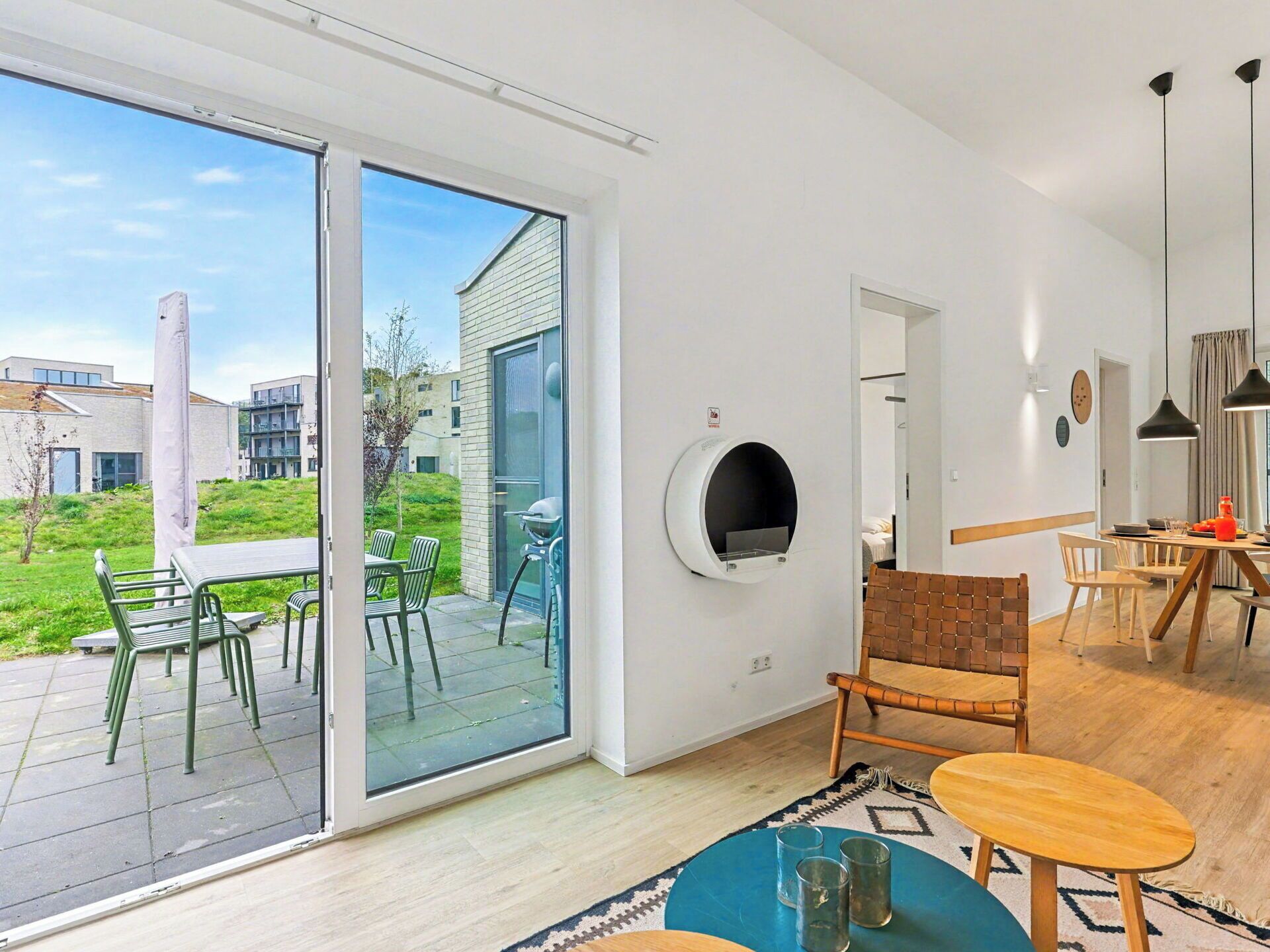 A modern living room with a sliding glass door leading to a patio, a built-in circular fireplace, and a dining area with a wooden table and chairs.