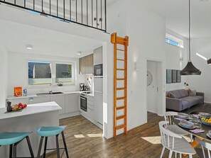 Eine moderne Küche mit einer zentralen Kochinsel, ein Essbereich mit Tisch für vier Personen und ein Wohnzimmer mit einer Couch.