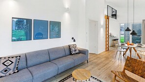 Ein modernes Wohnzimmer mit grauem Sofa, einem runden Couchtisch und einem Essbereich mit Holztisch und Stühlen. An der Wand hängen drei gerahmte Kunstwerke, im Hintergrund ist eine Treppe zu sehen.