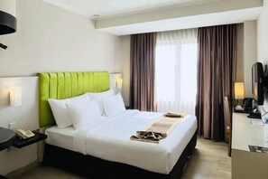 Een hotelkamer met een groen bekleden hoofdeinde, een witte spreidek, een nachtkastje met een lamp en een bureau met een computer en een telefoon.