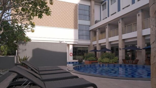 A pool area with lounge chairs and umbrellas, surrounded by a building with multiple balconies and a tiled floor. - Vrata Hotel Yogyakarta (Kalasan)