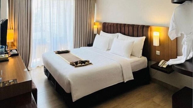 A hotel room with a large bed, a desk, a lamp, and a view of the city through the window. - Vrata Hotel Yogyakarta (Kalasan)