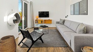 Un salon moderne avec un canapé gris, un fauteuil noir, une table basse en verre, un téléviseur à écran plat et, au mur, des tableaux encadrés représentant des paysages urbains.
