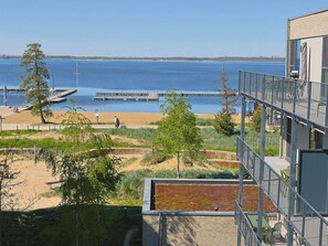 Blick auf einen Strand mit einem Steg, ein Gebäude mit Balkon und Windkraftanlagen in der Ferne.