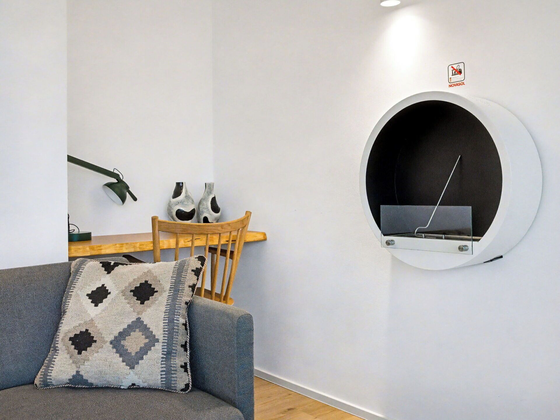 Un salon moderne avec une étagère ronde fixée au mur, une chaise en bois, un canapé gris agrémenté d’un coussin à motifs et une petite table sur laquelle sont posés deux vases.