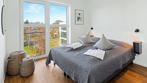 Un dormitorio con cama, almohadas, una mesita de noche, lámpara, un cuadro en la pared y vistas a través de un amplio ventanal.
