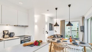 Ett modernt kök med vita skåp, en inbyggd ugn och en kaffebryggare. Det finns en matplats med ett träbord, stolar och utsikt över balkongen.