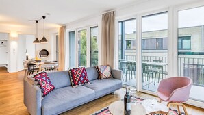 Een moderne woonkamer met een grijze bank, een ronde salontafel en een roze fauteuil. Er is een eethoek met een tafel en stoelen, en grote ramen met uitzicht op buiten.