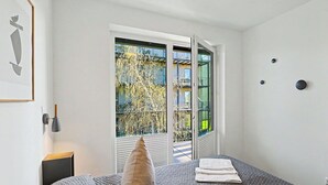Ein Schlafzimmer mit Bett, Kissen, Nachttisch, einem gerahmten Bild und einem Fenster mit Blick auf Gebäude und Bäume.