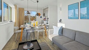 Ein modernes Wohnzimmer mit grauem Sofa, Essbereich mit Tisch und Stühlen, Küche im Hintergrund und großen Fenstern.