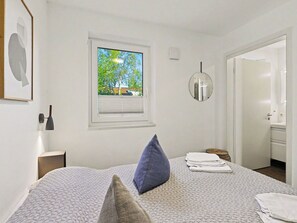 Ein Schlafzimmer mit einem Bett, einem Fenster mit Jalousien, einer Nachttischlampe, einem runden Spiegel und einer Tür zu einem Badezimmer.
