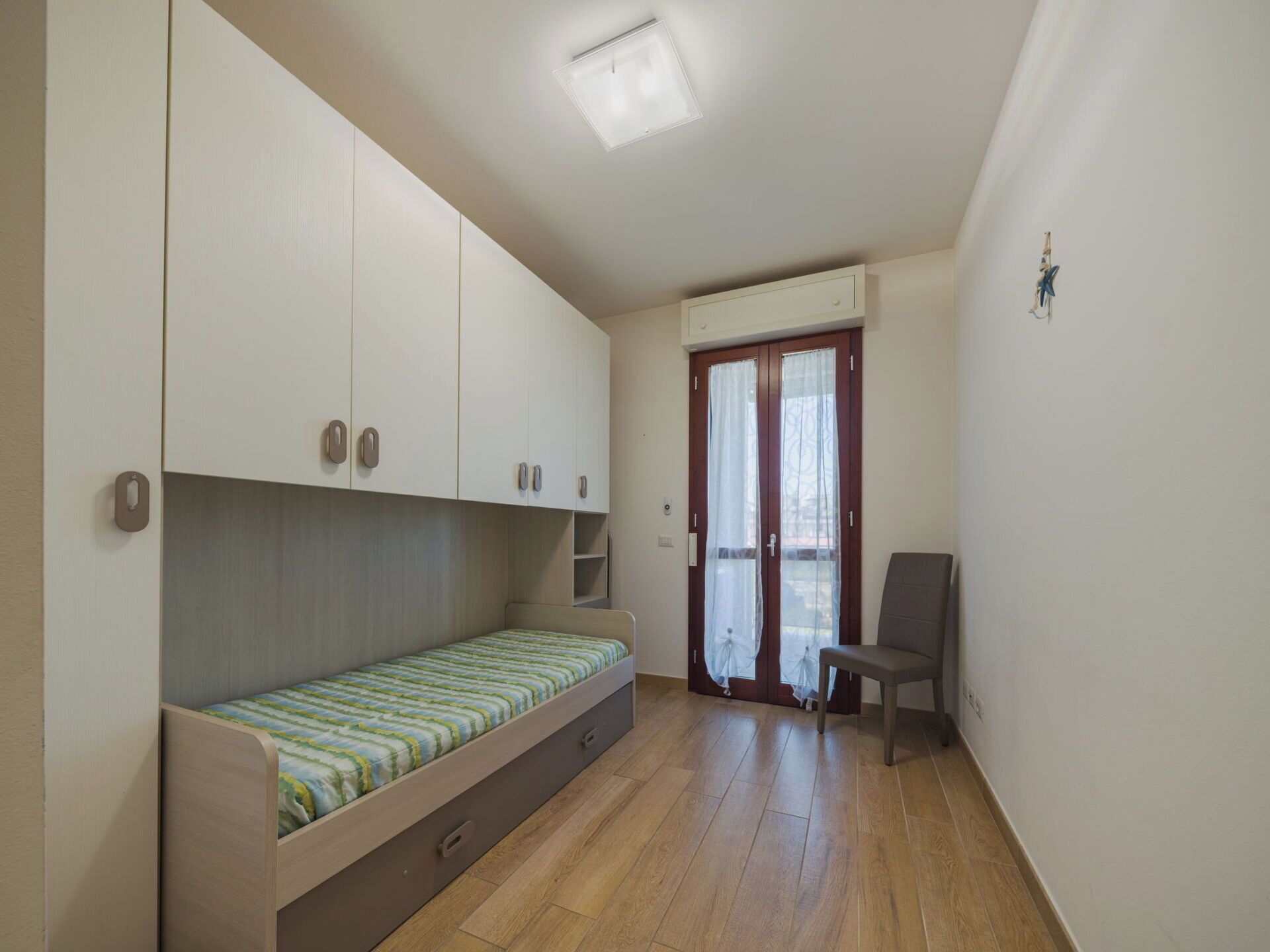 Een kleine, compacte kamer met een enkel bed, ingebouwde opbergruimte, een stoel en een deur met een raam.