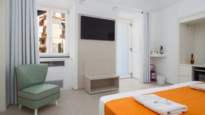 Habitación de hotel con cama, una silla, televisor, ventana con cortinas, extintor de incendios y un pequeño estante con artículos de aseo.