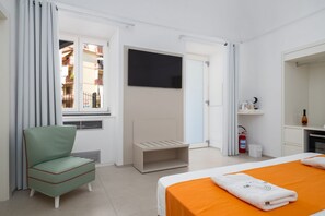 Ein Hotelzimmer mit einem Bett, einem Sessel, einem Fernseher, einem Fenster mit Vorhängen, einem Feuerlöscher und einer kleinen Ablage mit Pflegeprodukten.