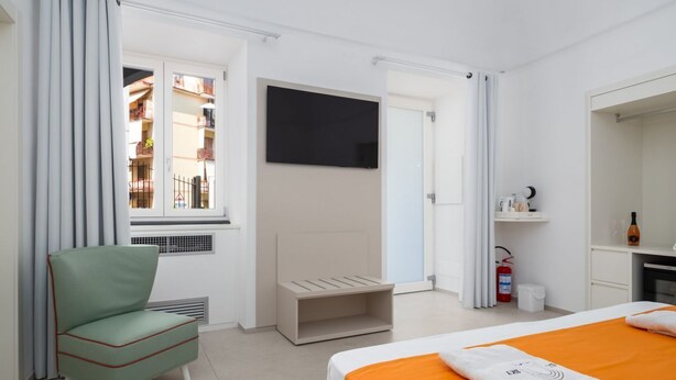 Habitación de hotel con cama, una silla, televisor, ventana con cortinas, extintor de incendios y un pequeño estante con artículos de aseo.